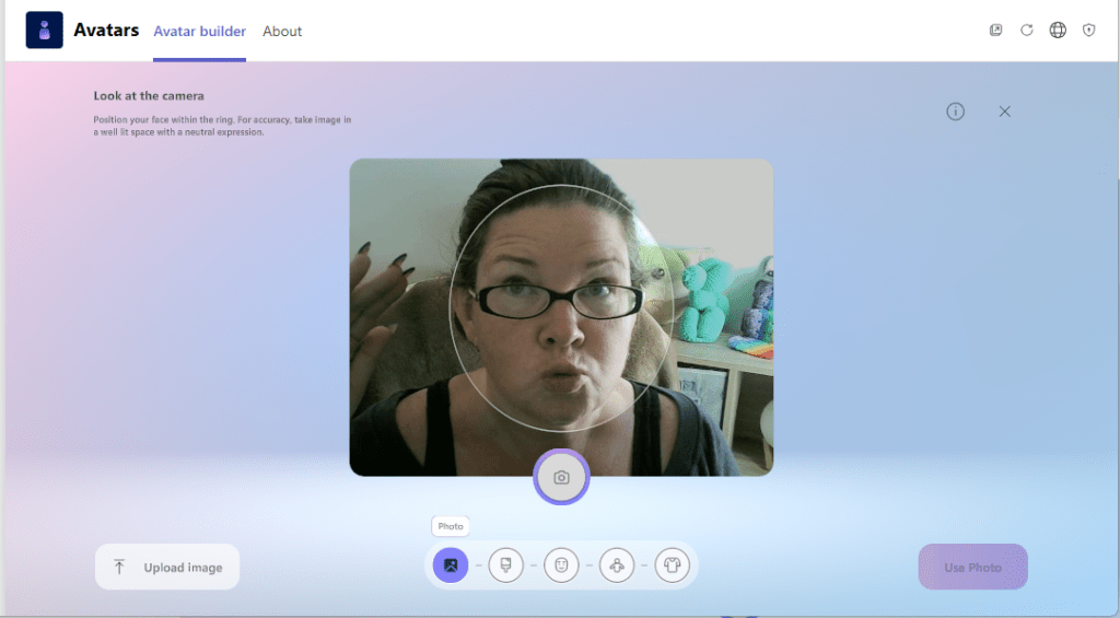 The image shows the "Avatar builder" section of the "Avatars" app within Microsoft Teams, focused on the camera interface for creating an avatar. The interface is divided into several sections:

1. **Header**: The top of the screen has three tabs - "Avatars," "Avatar builder" (selected), and "About." 

2. **Instructions**: Below the header, on the left side, there is a text instruction that reads, "Look at the camera. Position your face within the ring. For accuracy, take image in a well-lit space with a neutral expression."

3. **Camera View**: The central part of the screen shows a live camera feed with a person looking at the camera. The person is positioned within a circular frame guideline. The background features a home setting with stuffed toys and a lamp visible.

4. **Controls**: Below the camera view, there are several controls:
   - An "Upload image" button on the left.
   - A central camera icon within a circle to take the photo.
   - On the right, a purple "Use Photo" button.

5. **Additional Icons**: Below the camera button, there are smaller icons indicating different features or adjustments that can be made, including profile, gender selection, facial hair, accessories, and clothing options.

6. **Background**: The overall background features a gradient of light blue and pink, providing a clean and modern look to the interface. 

The interface is designed to guide users through the process of taking a suitable photo for creating an avatar.